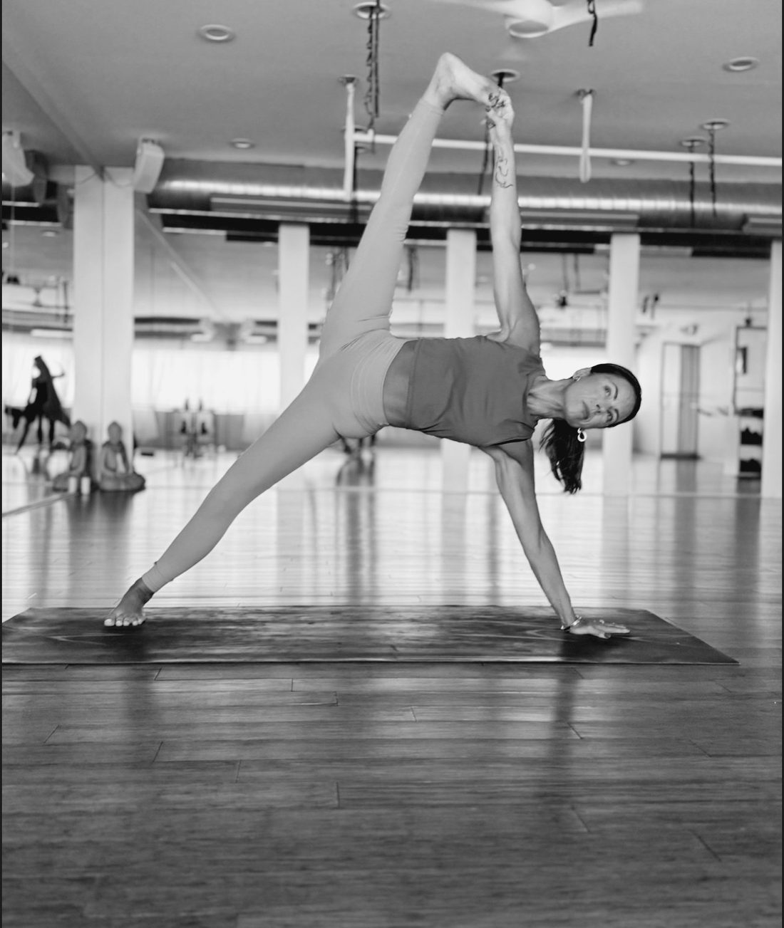 A teacher in side plank holding her raised foot, the studio's long room behind her.