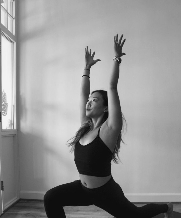 A teacher in a high crescent lunge by the studio's tall window, arms raised to the light.