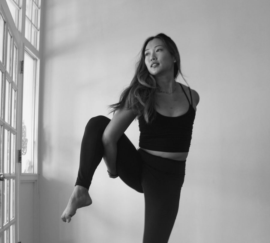 A teacher balancing on one leg, knee lifted, beside the studio's window.